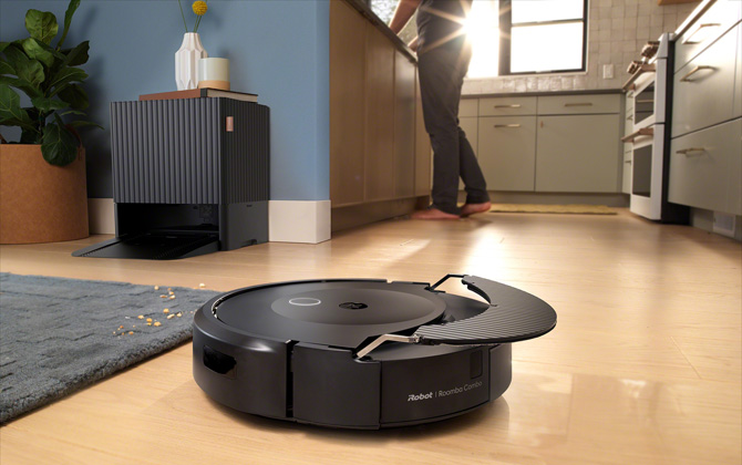 iRobot Roomba Comba 10 Max Robot + AutoWash Dock is shown in a living room. The mop pad is being lifted out of the way as the robot approaches carpeting. You can see the dock in the background with a vase, book, and mug on top. 