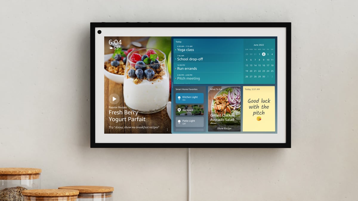 The Amazon Echo Show 15 is shown mounted on a wall in a kitchen. You can see a family calendar, a note, a couple of recipies, and smart home controls.